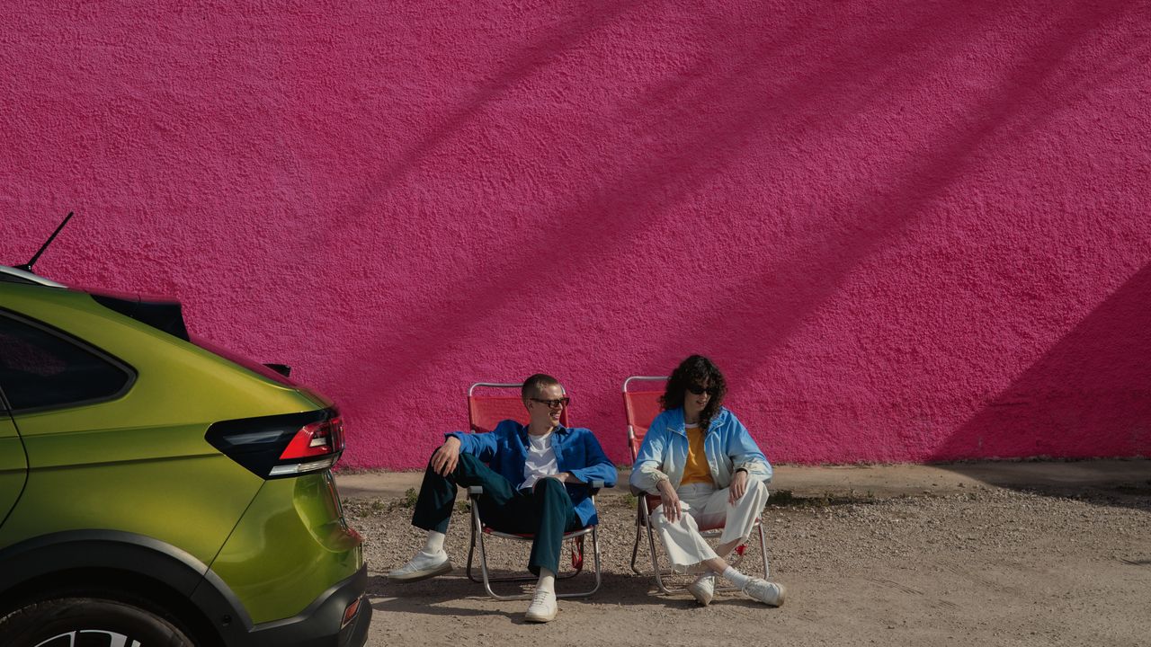 VW Taigo Heck in Grün, dahinter sitzen zwei Personen auf Campingstühlenht auf einem Parkplatz, ein Mann steht davor und blickt auf die Motorhaube.