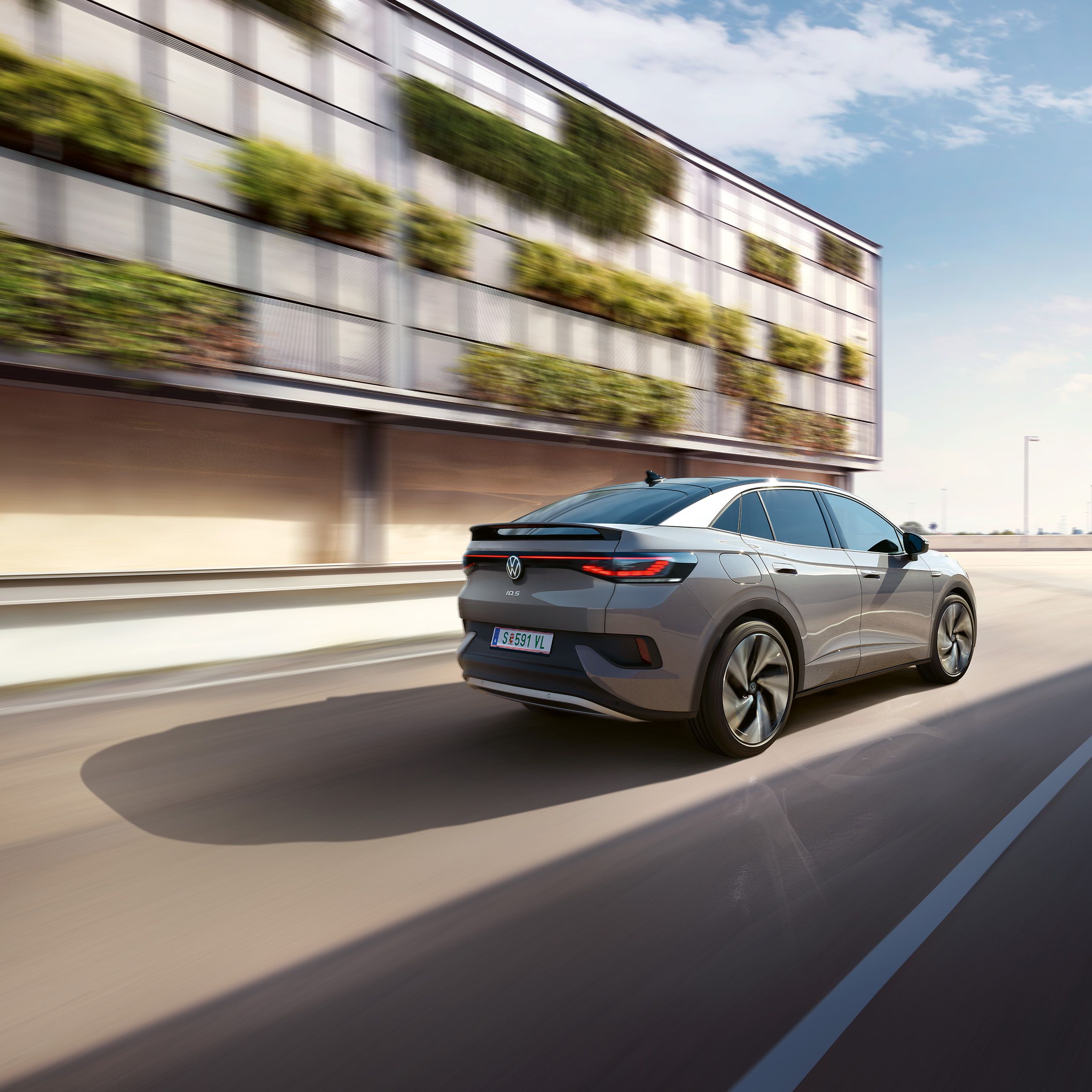 Fahraufnahme eines grauen VW ID.5 von schräg hinten, links ein helles, teilweise grün bewachsenes Gebäude