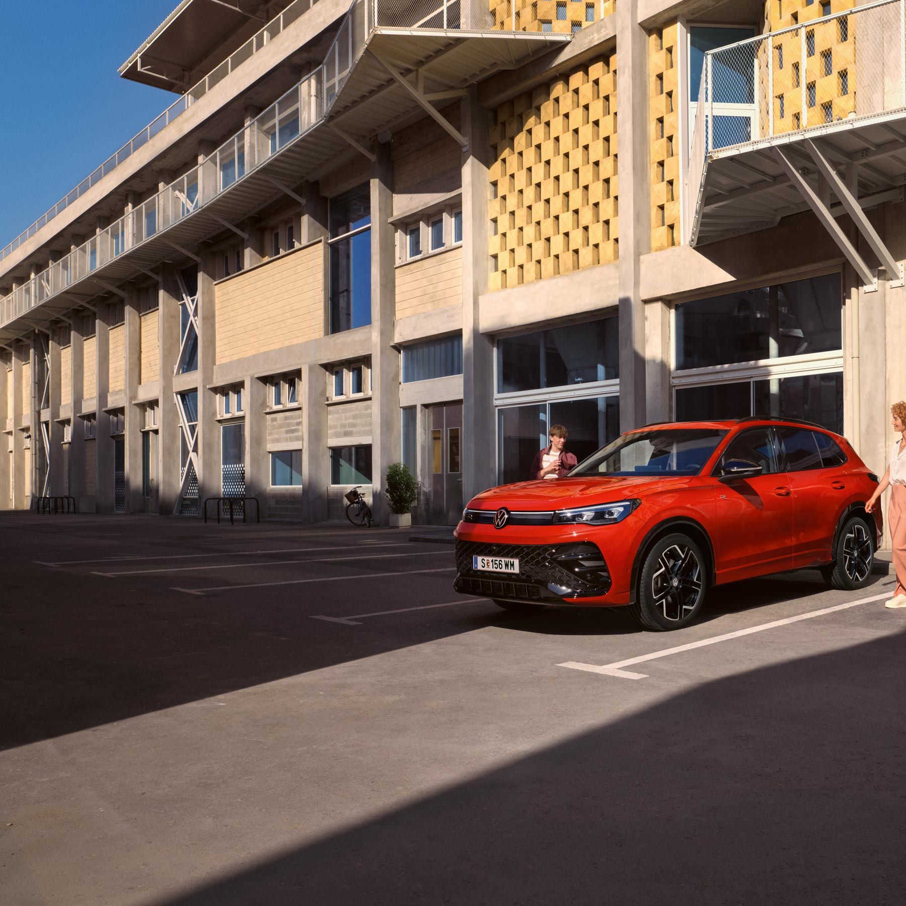Roter VW Tiguan in Seitenansicht steht vor einem Gebäude, eine junge Frau geht auf der Fahrerseite vorbei.