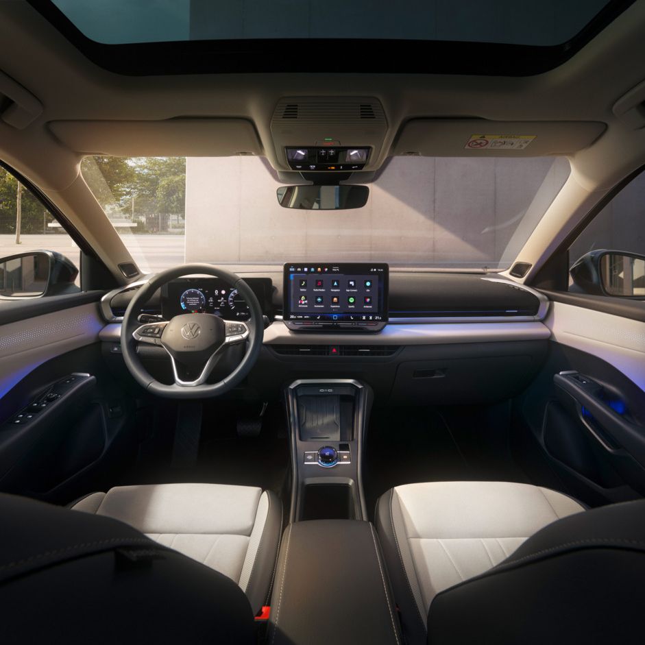 Das Bild zeigt das Innere eines T-Roc mit einem Sonnen- oder Glasdach. Das Cockpit hat ein großes Bildschirm-Display und ein Lenkrad mit mehreren Funktionen. Außen ist eine Grünfläche und ein Gebäude zu erkennen.