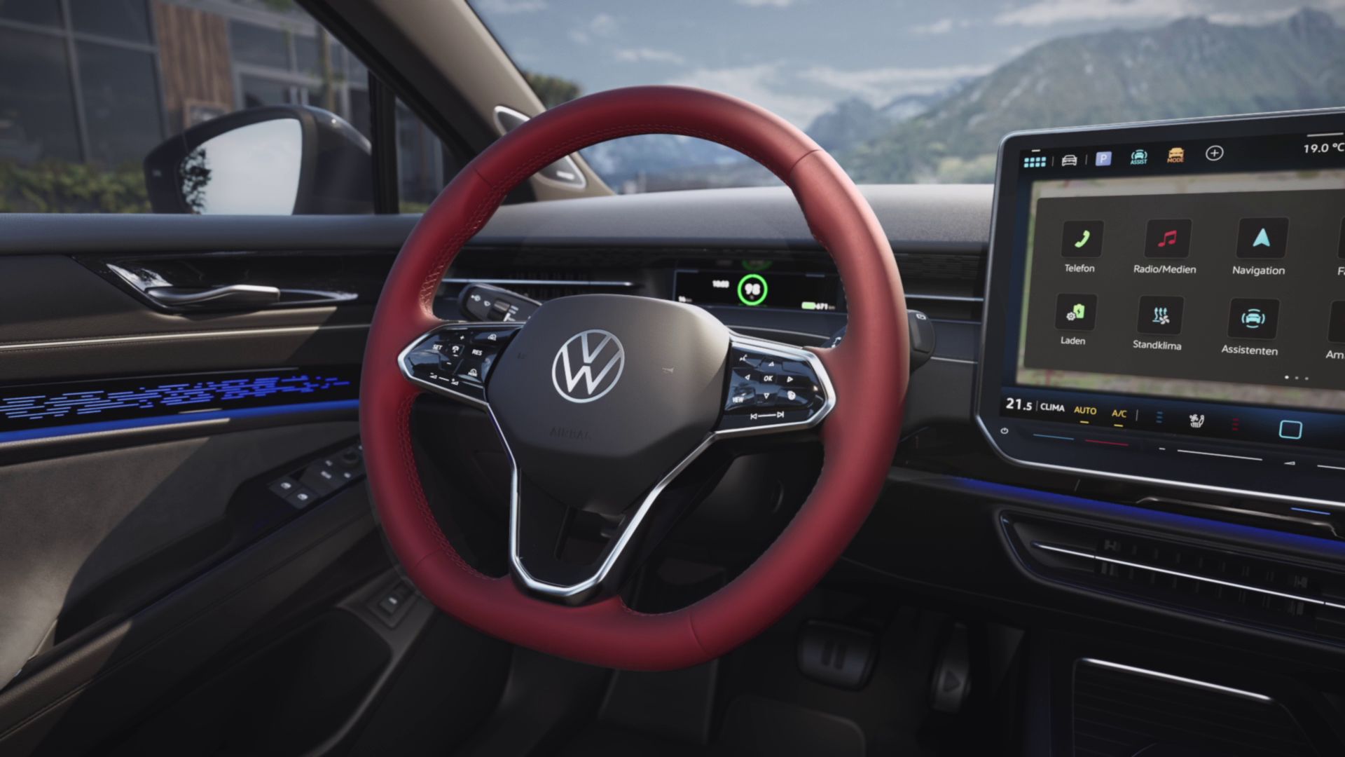 Das Bild zeigt den Innenraum eines modernen Autos. Im Vordergrund sieht man ein Lenkrad mit rotem Bezug und dem VW-Logo, rechts daneben ist ein großes Display mit verschiedenen Menüpunkten. Im Hintergrund sind Berge und eine Straße sichtbar.