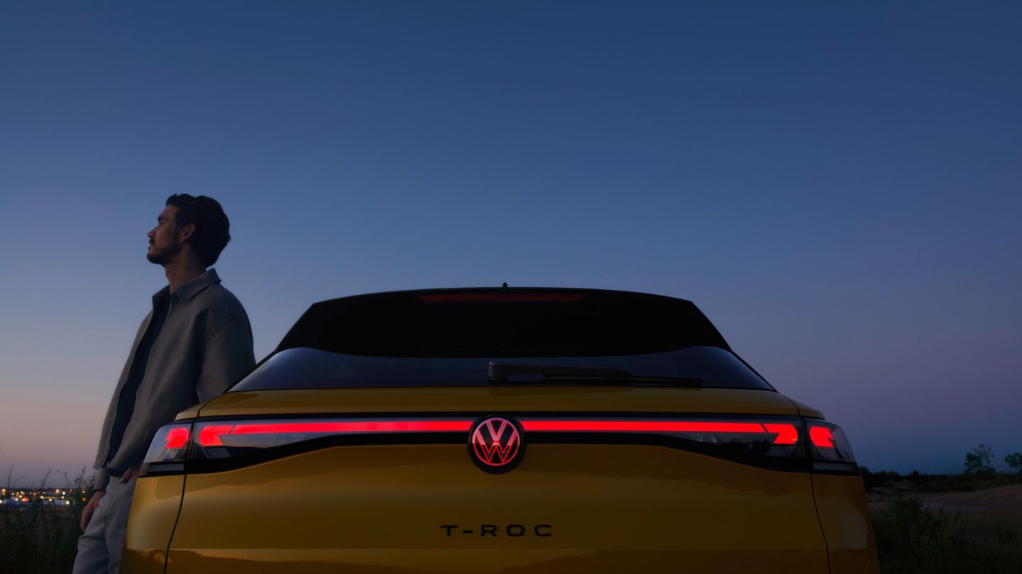 Ein Mann neben einem VW T-Roc R-Line mit eingeschalteten 3D-LED-Rückleuchten und rot-leuchtendem Logo.