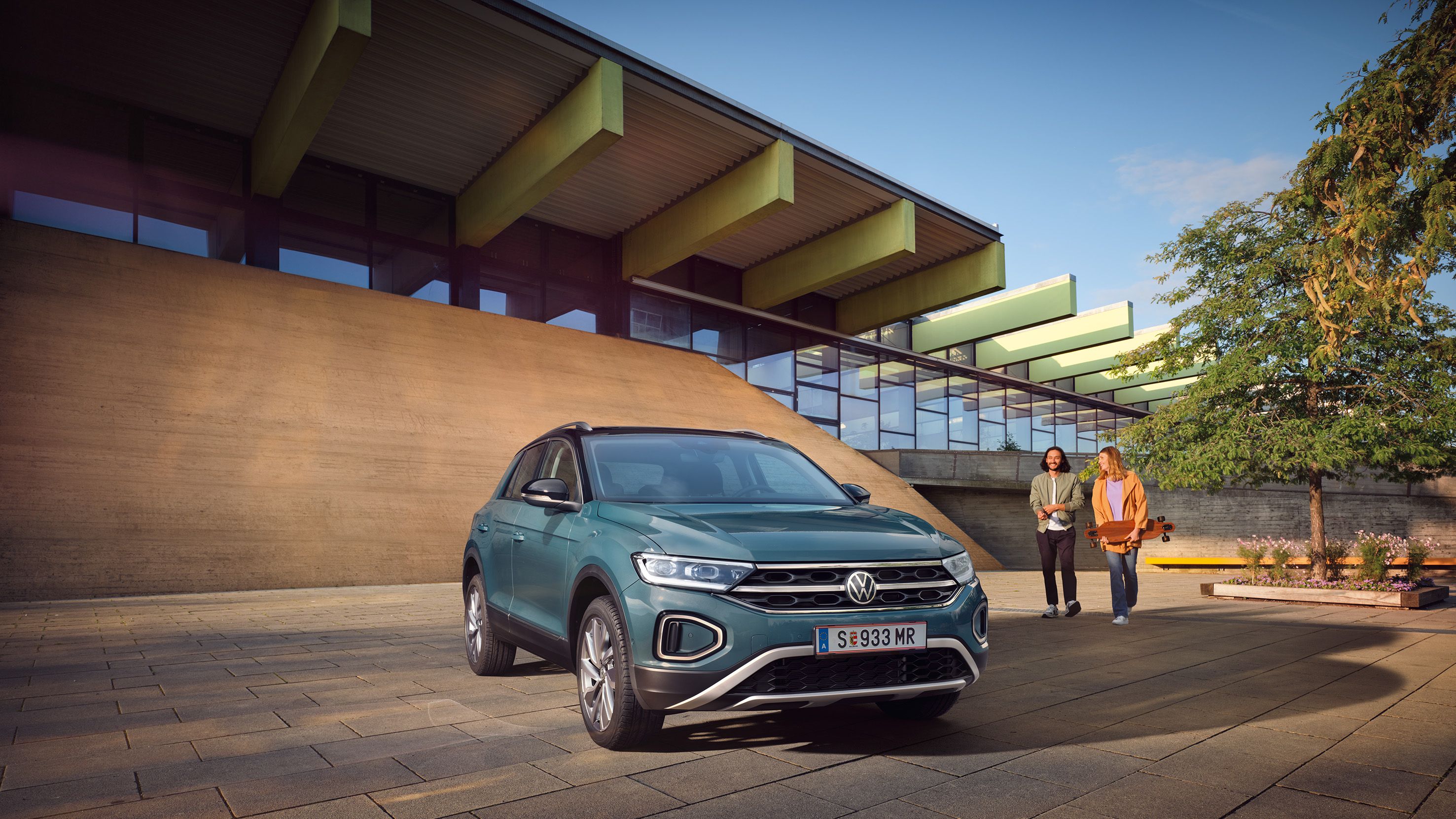 VW T-Roc Style in Blau vor Gebäude geparkt, Front und Seite sichtbar, 2 Personen gehen darauf zu.