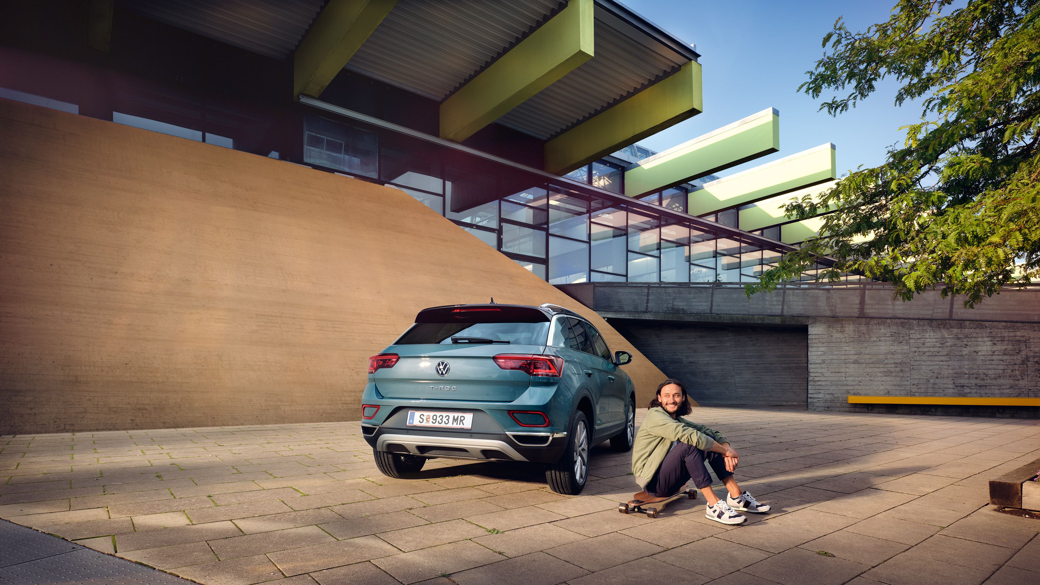 VW T-Roc Style in Blau vor Gebäude geparkt, Heck mit Rückleuchten sichtbar. Man sitzt auf Longboard neben dem Auto.