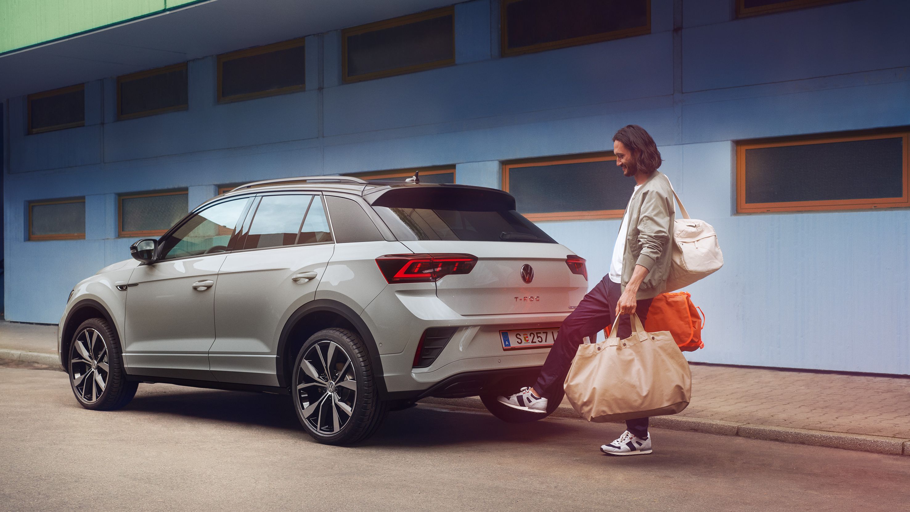 VW T-Roc R-Line in weiß vor einem Gebäude, Seite sichtbar, Mann steht hinter dem Auto und öffnet mit Fußschwenk die Kofferraumklappe