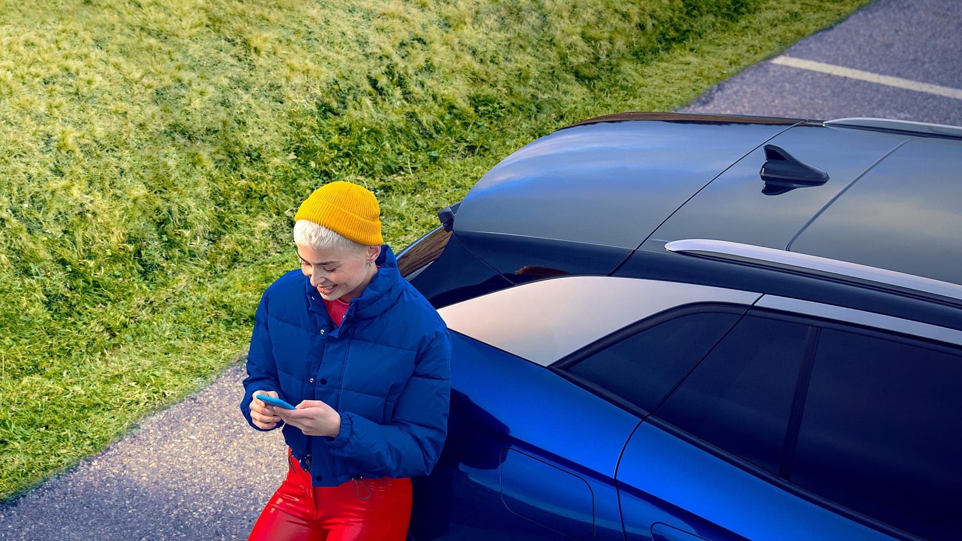 Heckansicht eines blauen VW ID.4 von schräg oben, sportlich gekleidete Person lehnt am Auto und bedient ein Smartphone