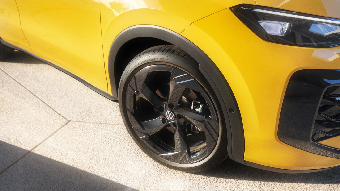 Das Bild zeigt einen Teil eines gelben T-Roc aus der Seitenansicht. Der Fokus liegt auf dem schwarzen Vorderrad und der Kotflügel. Das Auto steht auf hellen, rauen Bodenplatten.