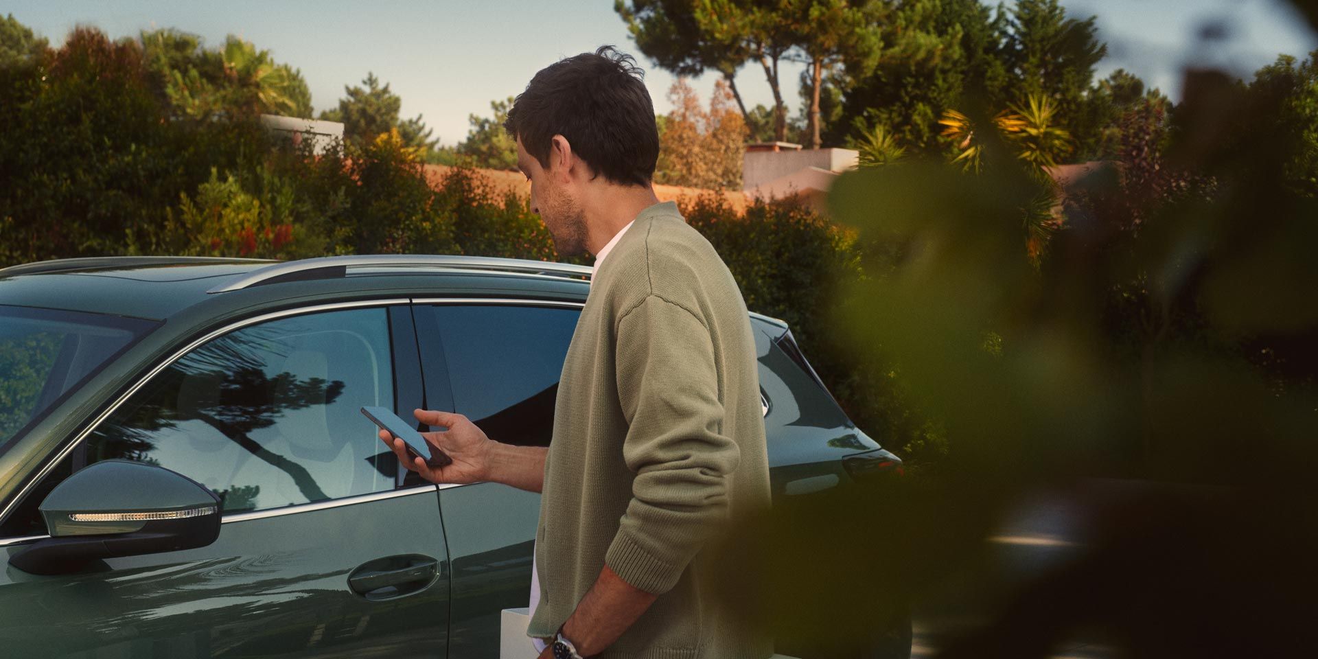 Ein junger Mann auf der Fahrerseite des dunkelgrünen VW Tiguan bedient den optionalen Park Assist Plus über sein Smartphone.
