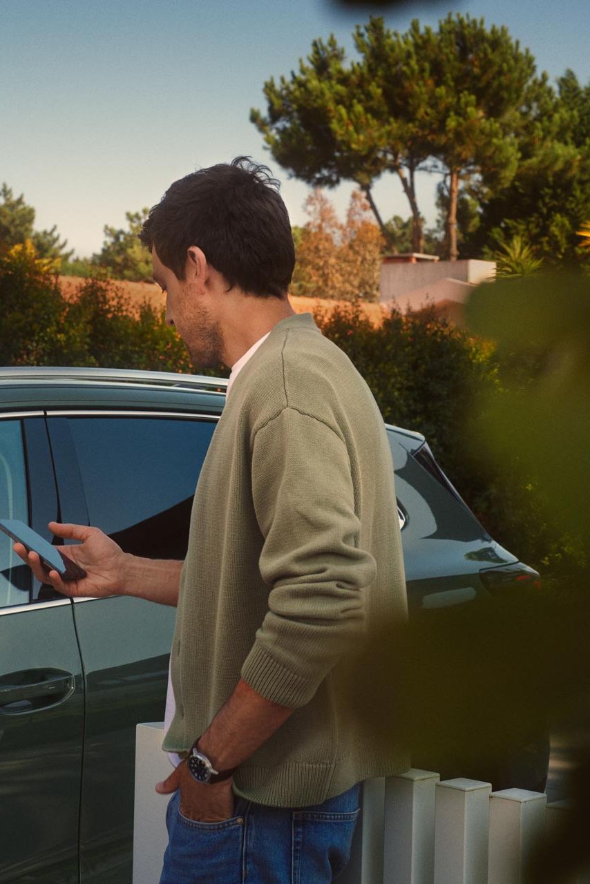 Ein junger Mann auf der Fahrerseite des dunkelgrünen VW Tiguan bedient den optionalen Park Assist Plus über sein Smartphone.